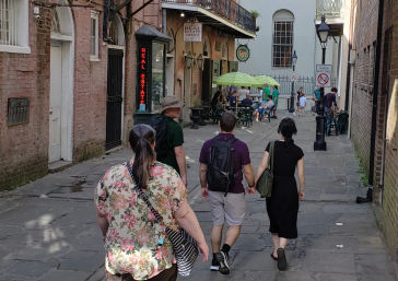 Sunlit narrow brick alley with cobblestone pavement, iron balcony, outdoor café with green umbrellas and people walking