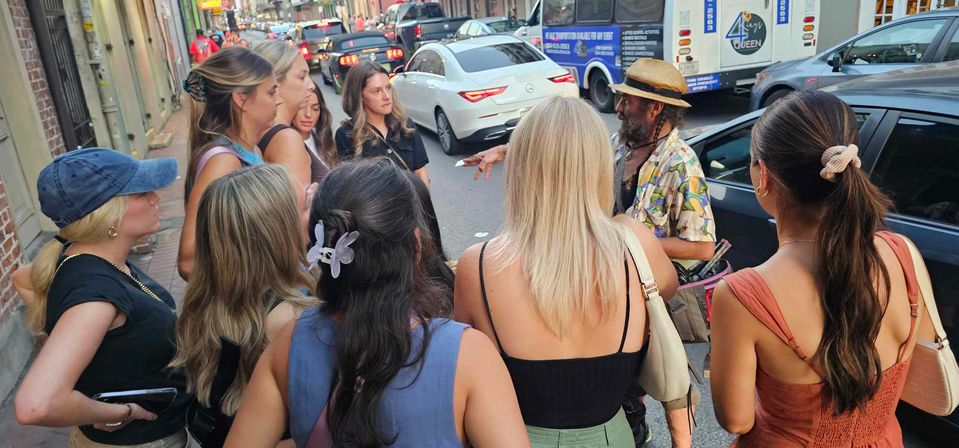 Group of young women gathered on a busy city street listening to a bearded street vendor in a straw hat and colorful shirt, with cars and a bus in the background.