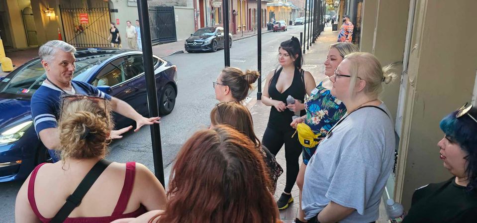Animated tour guide gesturing to a group on a historic downtown sidewalk during an evening walking tour, with parked cars and brick buildings in the background.