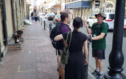 Tour guide in a sun hat speaking to a small group of tourists on a brick sidewalk lined with historic storefronts and parked cars