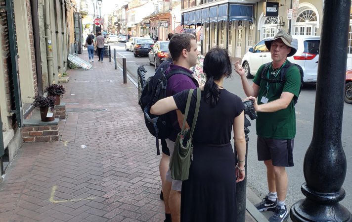Tour guide in a sun hat speaking to a small group of tourists on a brick sidewalk lined with historic storefronts and parked cars