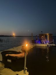 Cozy lakeside dock at dusk with tiki torches — a person soaking in a black portable hot tub while a small group relaxes by a wooden floating sauna lit with blue lights over calm water.