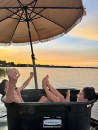 Two people relaxing in a black plastic tub on a dock under a fringed umbrella at a lakeside sunset, feet up over the water.