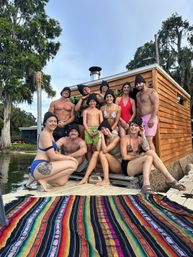 Smiling group of a dozen people in swimwear and black sauna hats posing on a lakeside dock beside a wooden floating sauna cabin, colorful woven blanket in the foreground and moss-draped cypress trees overhead.