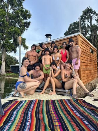 Smiling group of a dozen people in swimwear and black sauna hats posing on a lakeside dock beside a wooden floating sauna cabin, colorful woven blanket in the foreground and moss-draped cypress trees overhead.