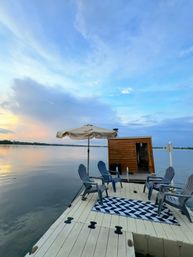 Lake dock at sunset with calm water, four Adirondack chairs, geometric rug, beige umbrella and a small wooden lakeside cabin/sauna under a soft blue sky