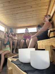 Group of five people relaxing in a wood‑paneled sauna with natural light streaming through windows, wearing sauna hats and seated around white crystal singing bowls for a meditative sound‑bath wellness session.