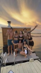Seven friends in swimwear and sauna hats posing and waving on a floating wooden dock beside a small sauna cabin at a lake sunset with a golden sky.