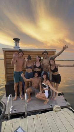 Seven friends in swimwear and sauna hats posing and waving on a floating wooden dock beside a small sauna cabin at a lake sunset with a golden sky.