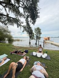 Lakefront outdoor sound bath with people lying on yoga mats under Spanish-moss oak trees, a practitioner seated with crystal singing bowls on a mat, and a wooden dock extending into a calm lake