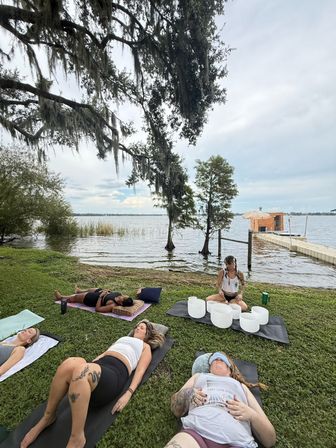 Lakefront outdoor sound bath with people lying on yoga mats under Spanish-moss oak trees, a practitioner seated with crystal singing bowls on a mat, and a wooden dock extending into a calm lake