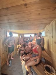 Group of people enjoying a cozy wooden sauna interior, seated on tiered benches in swimwear and wool sauna hats with sunlight streaming through narrow windows and a wooden bucket on the floor.