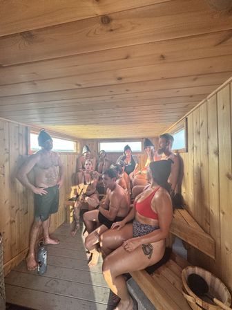 Group of people enjoying a cozy wooden sauna interior, seated on tiered benches in swimwear and wool sauna hats with sunlight streaming through narrow windows and a wooden bucket on the floor.