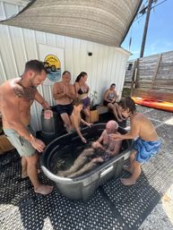 Multi‑generation group laughing around an outdoor cold‑plunge tub under a shade sail in sunny Florida, kids splashing an elderly man while adults watch near a wooden fence and kayak