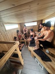 Group of adults relaxing and chatting in a traditional wooden sauna with bench seating and black sauna hats, sweating and enjoying a lake view through narrow windows.