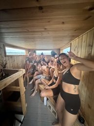 Wood-paneled lakeside sauna interior with a smiling group in swimwear and sauna hats on benches, low windows revealing a calm lake view — playful wellness moment.