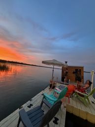 Colorful Adirondack chairs and umbrella on a lakeside dock at sunset, people relaxing near a small wooden shed with orange sky reflecting on calm water