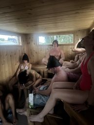 Group of people relaxing in a small wood‑paneled indoor sauna, wearing swimsuits and black felt sauna hats while seated on tiered wooden benches beneath narrow windows