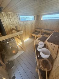 Cozy lakeside wood‑paneled sauna interior with tiered benches and black mats, wood‑burning stone heater, wooden ladles and white crystal singing bowls, narrow windows framing a calm lake view