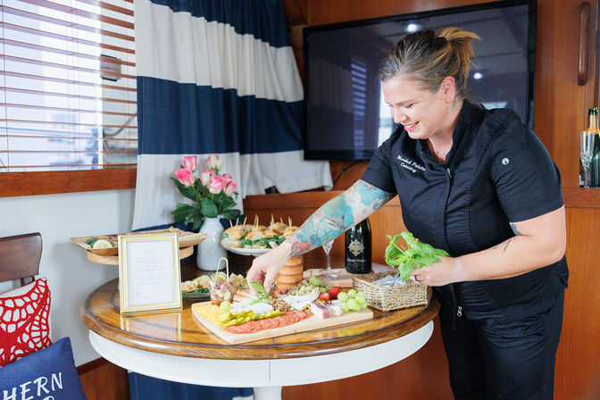 Chef arranging colorful charcuterie board with cheeses, grapes, cured meats and fresh herbs on a round table inside a yacht cabin, part of a catered spread.