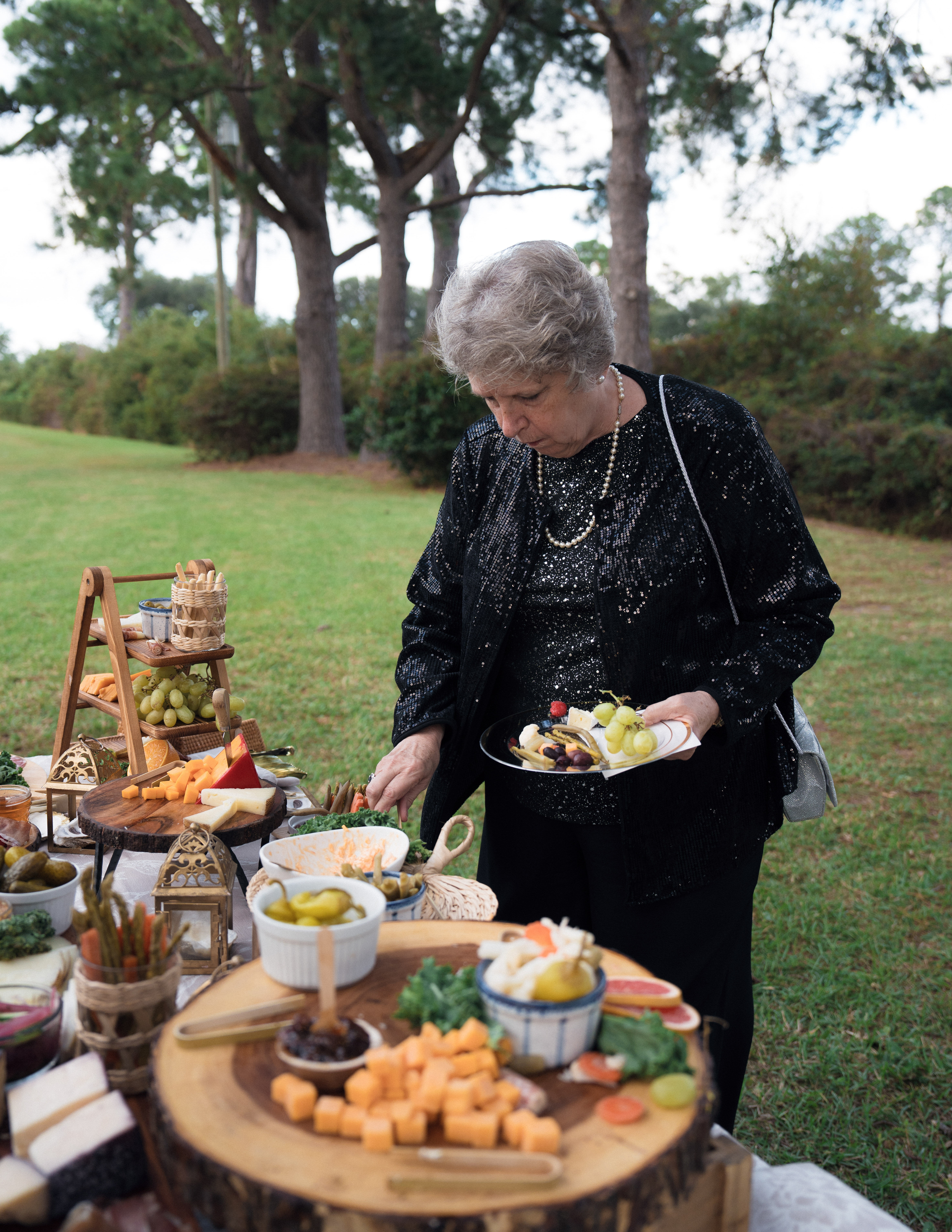 Older woman in a sparkly black jacket serving herself from a rustic outdoor charcuterie and cheese spread on wooden slabs, with grapes and pickles on a grassy lawn framed by pine trees.