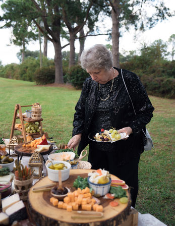 Older woman in a sparkly black jacket serving herself from a rustic outdoor charcuterie and cheese spread on wooden slabs, with grapes and pickles on a grassy lawn framed by pine trees.