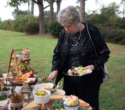 Older woman in a sparkly black jacket serving herself from a rustic outdoor charcuterie and cheese spread on wooden slabs, with grapes and pickles on a grassy lawn framed by pine trees.