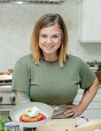 Smiling home cook in a green T‑shirt and striped apron presents a plated golden fritter on bright beet purée topped with a dollop of cream in a modern home kitchen