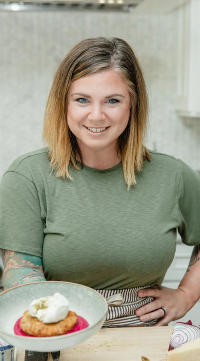 Smiling home cook in a green T‑shirt and striped apron presents a plated golden fritter on bright beet purée topped with a dollop of cream in a modern home kitchen