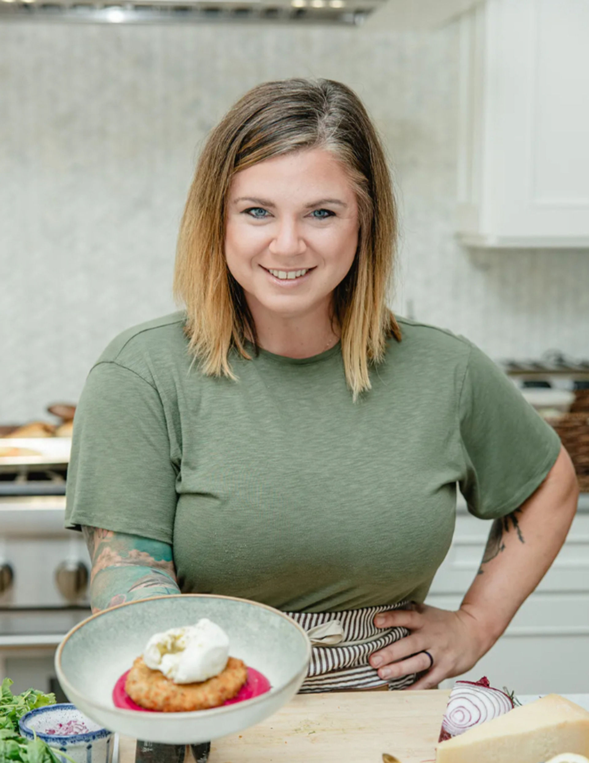 Smiling home cook in a green T‑shirt and striped apron presents a plated golden fritter on bright beet purée topped with a dollop of cream in a modern home kitchen