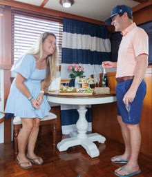 Smiling woman in a pale blue dress laughing while a man in a coral polo pops champagne beside a round table with appetizers and pink roses inside a yacht cabin.