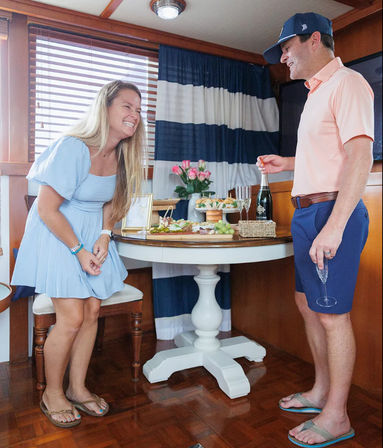 Smiling woman in a pale blue dress laughing while a man in a coral polo pops champagne beside a round table with appetizers and pink roses inside a yacht cabin.