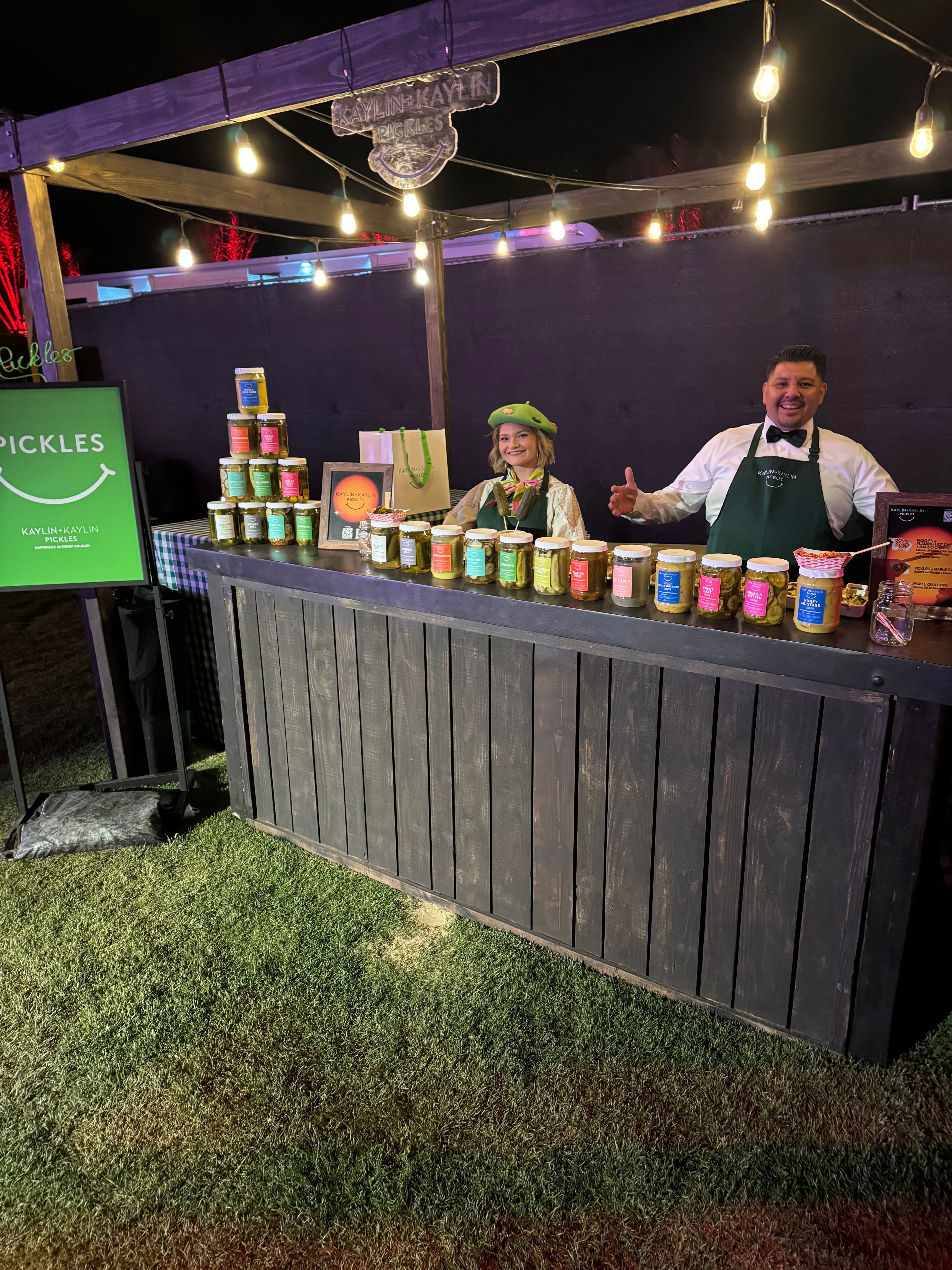 Night market pickle stall under warm string lights — two smiling vendors behind a rustic wooden counter displaying colorful jars of artisanal pickles at an outdoor festival on grass.