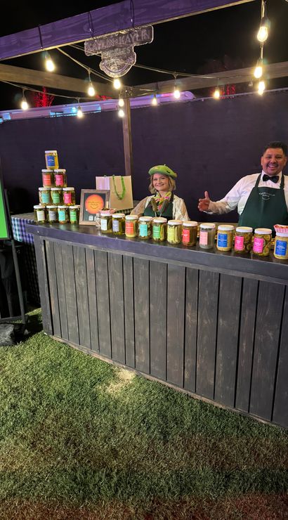 Night market pickle stall under warm string lights — two smiling vendors behind a rustic wooden counter displaying colorful jars of artisanal pickles at an outdoor festival on grass.