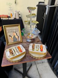 Outdoor pop-up pickle bar with a three-tiered pickle fountain, gourmet topped pickle bites on trays, and a framed sign on a wooden display table.