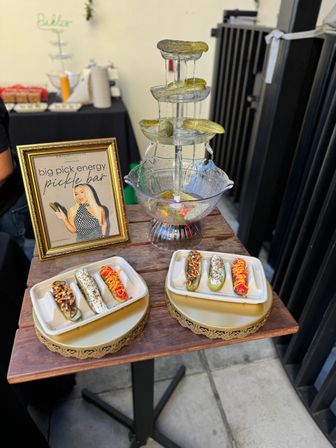 Outdoor pop-up pickle bar with a three-tiered pickle fountain, gourmet topped pickle bites on trays, and a framed sign on a wooden display table.