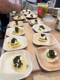 Close-up of caviar canapés: ridged potato chips topped with pickle slice, dollop of cream and black caviar served on small palm-leaf plates at a tasting counter.