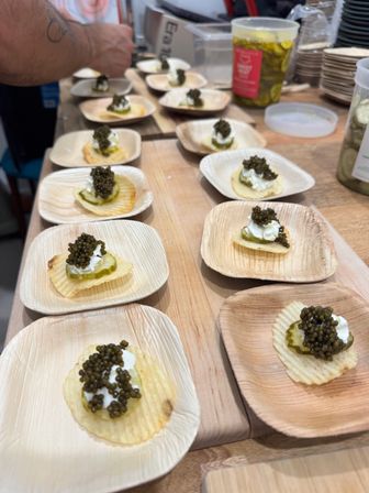 Close-up of caviar canapés: ridged potato chips topped with pickle slice, dollop of cream and black caviar served on small palm-leaf plates at a tasting counter.