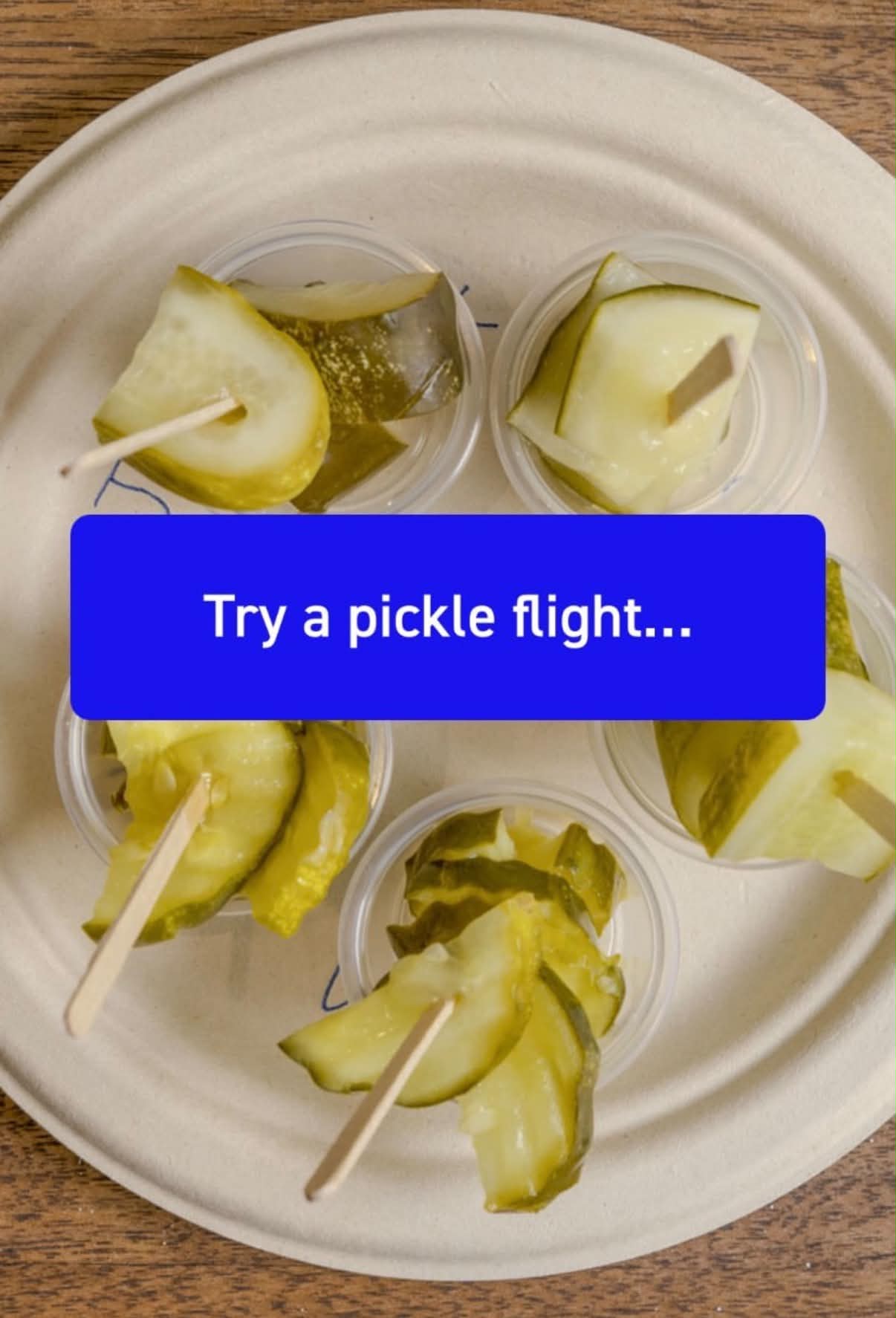 Top-down view of a pickle flight tasting sampler — small cups of dill pickle slices on toothpicks arranged on a compostable plate over a wooden table, with a blue label reading "Try a pickle flight...".