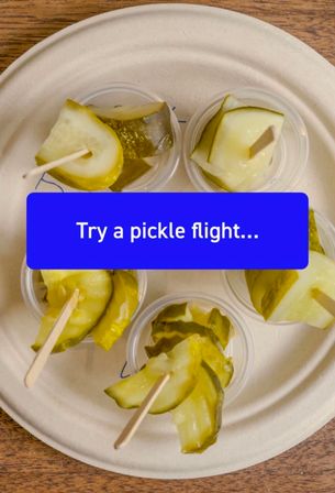 Top-down view of a pickle flight tasting sampler — small cups of dill pickle slices on toothpicks arranged on a compostable plate over a wooden table, with a blue label reading "Try a pickle flight...".