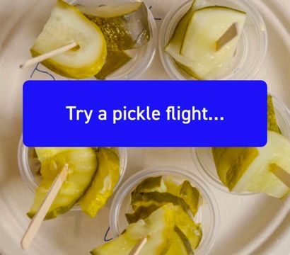 Top-down view of a pickle flight tasting sampler — small cups of dill pickle slices on toothpicks arranged on a compostable plate over a wooden table, with a blue label reading "Try a pickle flight...".