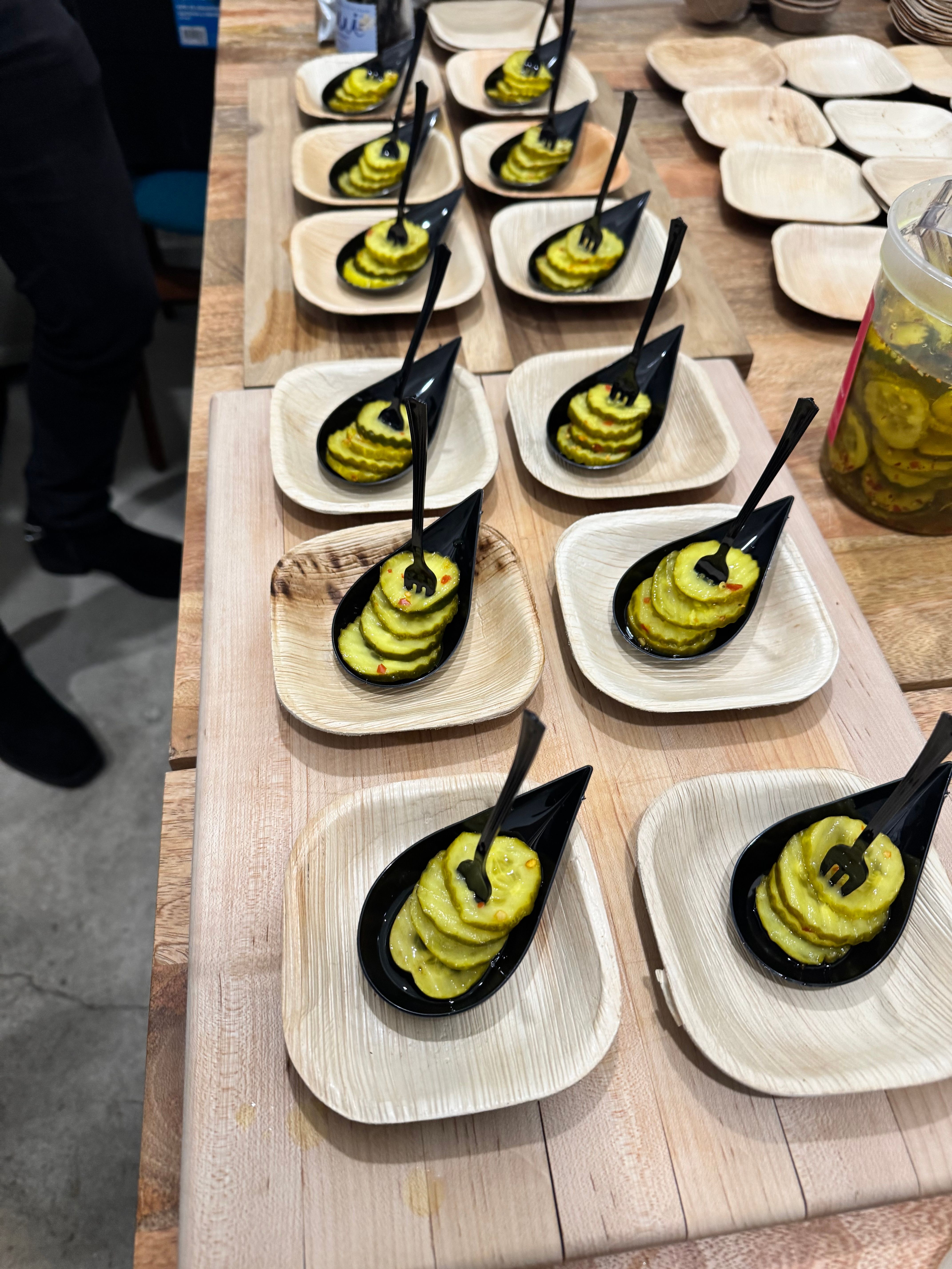 Stacked crinkle-cut pickle slices with mini forks in black tasting spoons, arranged on palm-leaf plates and wooden boards at a tasting station