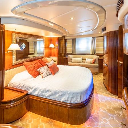 Luxurious wood‑paneled yacht master stateroom with curved ceiling, large bed in white linens and coral accent pillows, ornate wall mirror, cozy sofa and warm ambient lighting