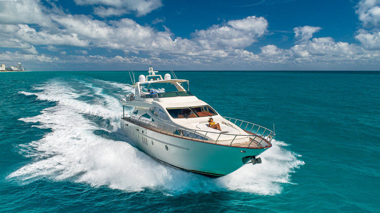 Luxury white motor yacht speeding through turquoise ocean with a trailing white wake, blue sky with fluffy clouds, person sunbathing on the bow and a distant coastline.