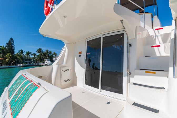 Luxury yacht stern deck with sliding glass door, curved white stairs to upper deck and mounted life ring, turquoise marina water and palm-tree lined shoreline under a clear blue sky.