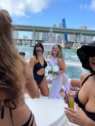 Group of women partying on a yacht in Miami harbor — swimsuits, a woman in white holding a bouquet, city skyline and raised bridge in the background on a sunny day.