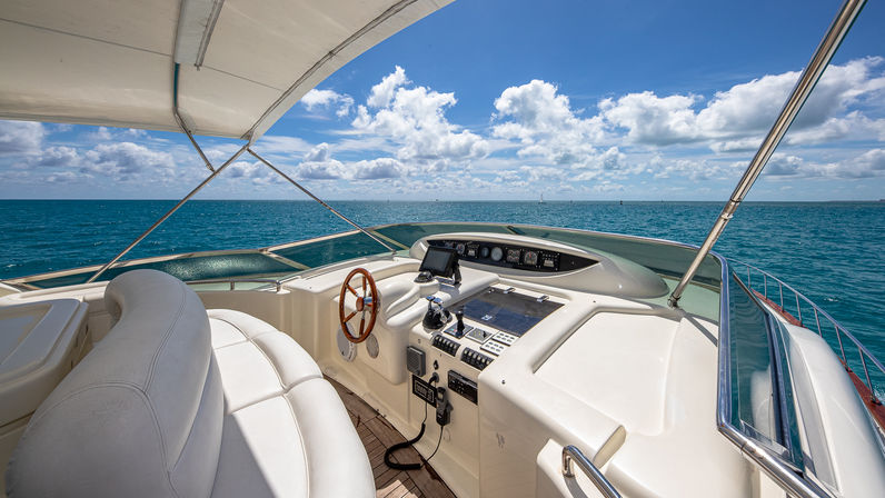 Sun-soaked luxury yacht helm with white leather seating, wooden steering wheel and navigation console under a canvas shade, turquoise sea and blue sky with fluffy clouds on the open ocean