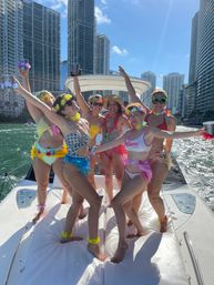 Six women in colorful swimsuits and leis partying on a yacht in Miami’s downtown waterfront with glass high-rises and a bright blue sky