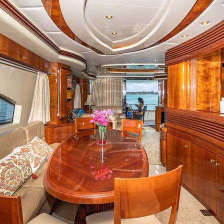 Luxury yacht interior — glossy wood-paneled salon with oval dining table topped with fresh pink flowers, cushioned seating, recessed ceiling lights and panoramic aft windows framing a sunny waterfront harbor view.