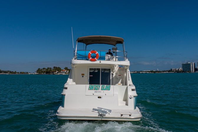 Rear view of a white motor yacht cutting through turquoise coastal waters under a bright blue sky, with an upper deck canopy, orange life ring, palm-lined shore and distant city skyline.